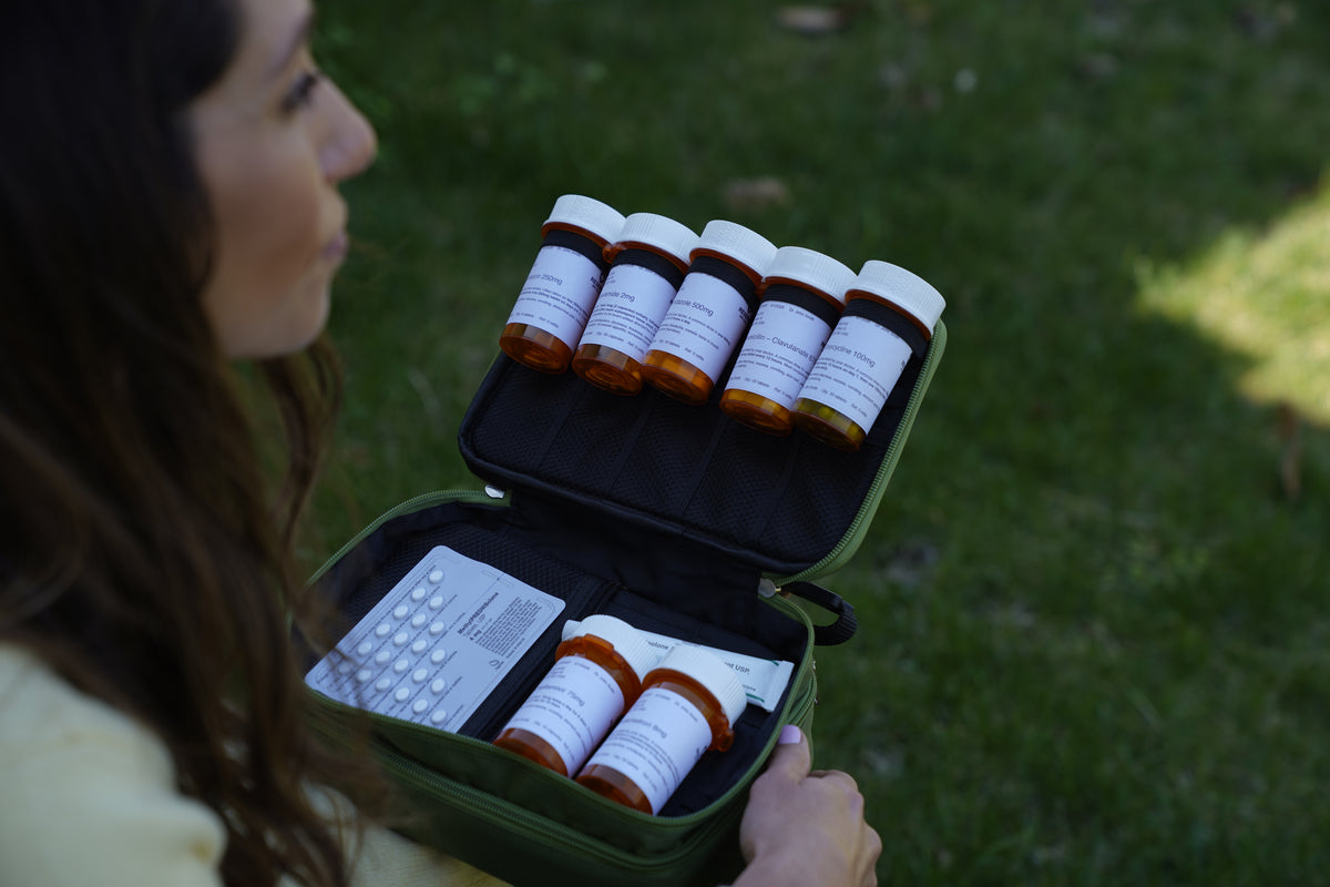 Over-the-shoulder view of a woman holding an open olive green Reserve Med Supply medical kit outdoors, showcasing organized rows of amber prescription medication bottles and a blister pack against a grassy background.