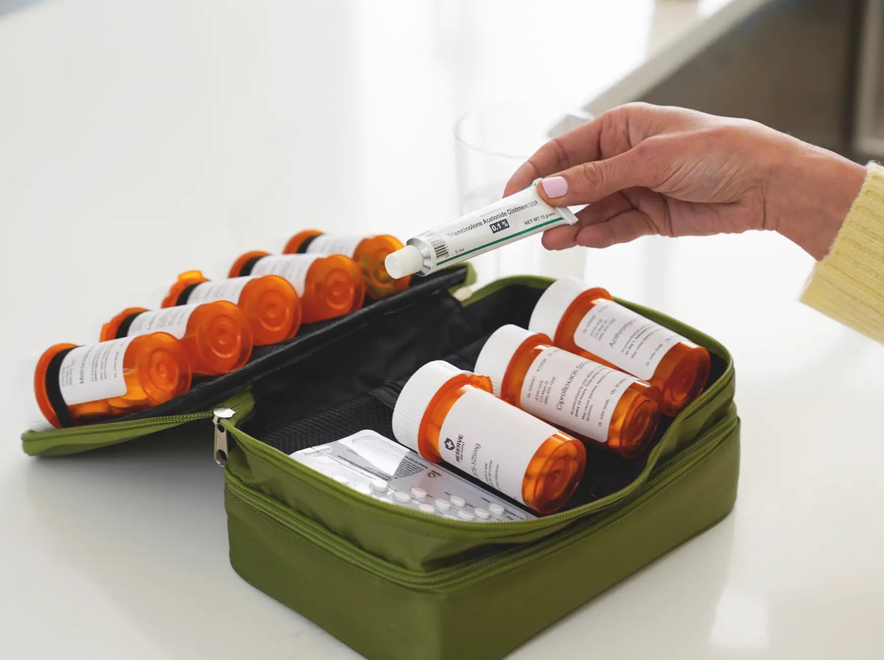 A person's hand holding a tube of Triamcinolone Acetonide ointment above an open olive green Reserve Med Supply case filled with several amber prescription pill bottles and a blister pack of medicine on a clean, white countertop.