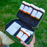 Over-the-shoulder view of an open olive green Reserve Med Supply medical kit outdoors, showcasing organized rows of amber prescription medication bottles and a blister pack against a grassy background.